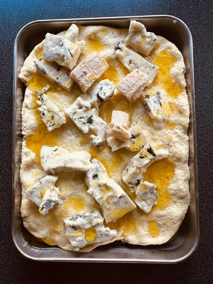 A rectangular focaccia topped with chunks of blue cheese and drizzled with olive oil is set on a baking tray.