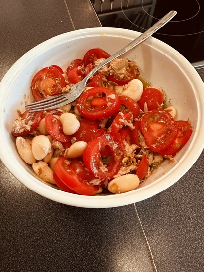 A bowl contains a salad made of sliced tomatoes, white beans, tuna, and dressing, with a fork placed on the side.