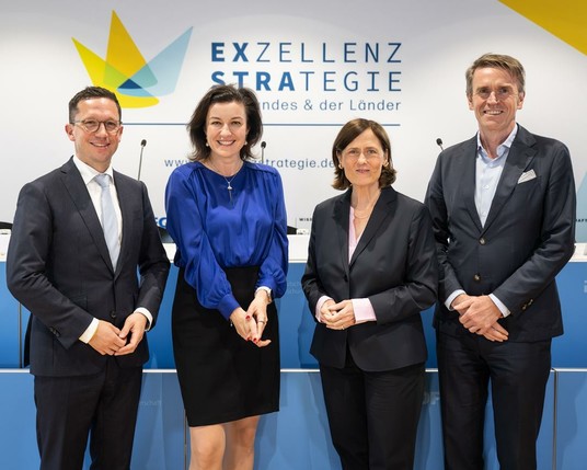 Vier Personen stehen vor einer Wand mit dem Schriftzug 'Exzellenzstrategie des Bundes und der Länder', wobei zwei Männer Anzüge tragen und zwei Frauen in Business-Outfits gekleidet sind.