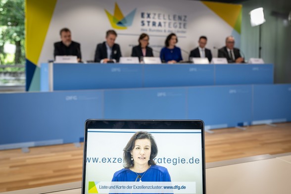 Im Vordergrund ein Bildschirm mit Bundesforschungsministerin Dorothee Bär, im Hintergrund ein Podium.