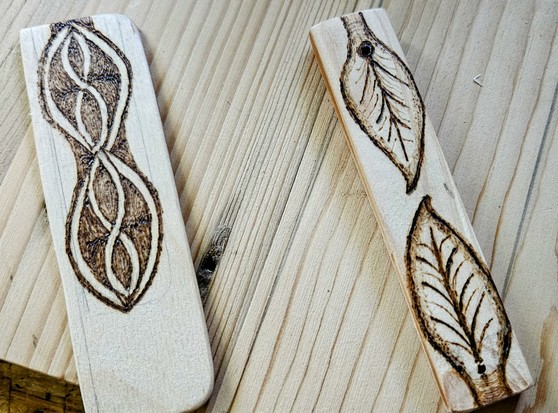 Two small, scrap pieces of wood. One has two leaf patterns burned onto it. The other are vertically aligned, eye-shaped loops, kinda knotting in the middle.