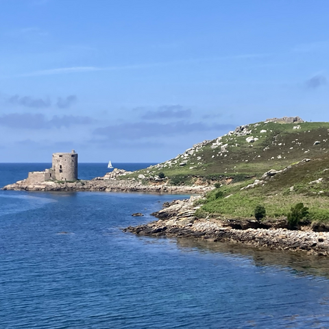 A view of Cromwell’s Castle on Tresco. It is a round stone castle jutting out on a low promontory. To the right is rocky shore and green hills dotted with Bronze Age gravestone cairns. In the left foreground and then beyond the castle there os clean blue sea.