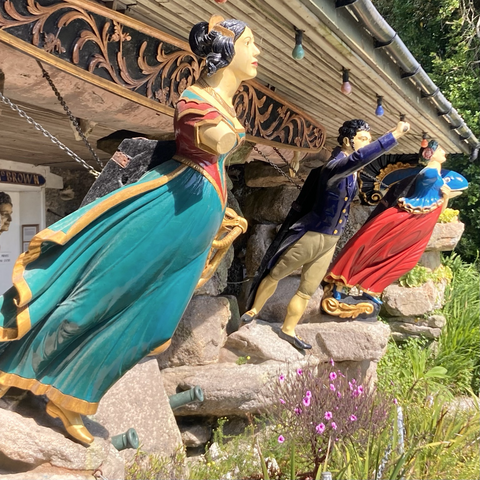 A row of ship figureheads from shipwrecks. They have been repainted/restored. Nearest the camera is a woman in a gold and turquoise 17th century outfit. Next to her is a man pointing forwards and up. He wears gold breeches and a navy jacket. Then there is a woman in a red dress ans blie top and finally a dolphin figurehead