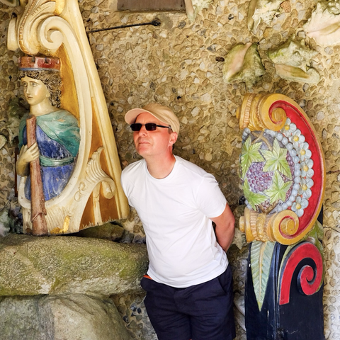 Me posing as a figurehead, leaning forward from the waist with my hands behind my back. I am wearing a beige cap, white t-shirt and navy shorts. I am in between a small female figurehead and a brightly painted unidentified crescent shaled part of a ship