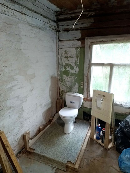 Same corner, with the toilet still standing but the flush reservoir slightly crooked. The cubicle is gone apart from the floor that the toilet is bolted to. There's a limewashed brick wall on one side and a wooden window in a log wall on the other. Part of the log wall has a green door panel nailed to it. On the side stands a quickly erected stand for a toilet paper roll and some other utensils, weighed down with a brick. Some bin bags collect various junk.
