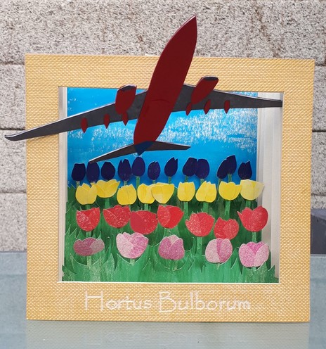 An accordion book (a pull-out diorama made of paper) with rows of pink, red, yellow and purple-black tulips cut out of paper under a clear blue sky. The flowers are heavily overshadowed by a large plane, cut out of cardboard, flying low overhead.