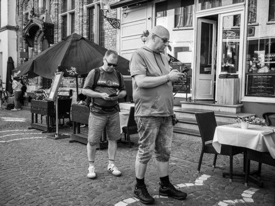The image is a black and white photograph depicting two men standing on a cobblestone street in front of a restaurant. The man in the foreground is bald, wearing glasses, a short-sleeved shirt, and knee-length shorts, and is focused on his smartphone. The man in the background is wearing a t-shirt, shorts, and sneakers, and is also looking at his phone.
