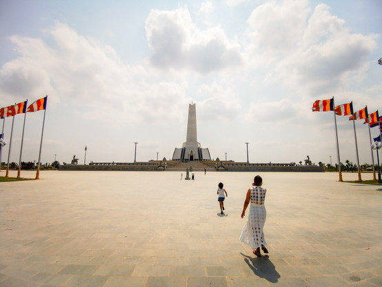Photo shows a girl running towards a tall stele monument. The entry to the area is flanked by red, white and blue Cambodian flags, and the five banded Buddhist flag. Her mother is walking after her child. It is a sunny day with partly cloudy blue sky. The area in front of the monument are light grey stone. The monument itself is even lighter.