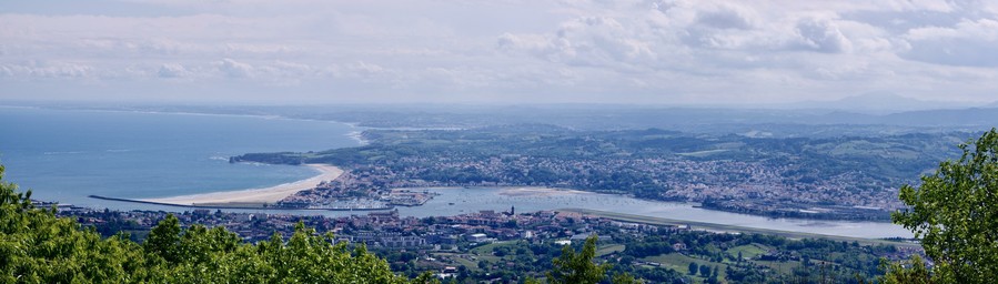 A panoramic view of a coastal landscape, featuring a sandy beach, a river leading to the ocean, and a town with various buildings and greenery in the foreground. The sky is partially cloudy, and the horizon features rolling hills in the distance.