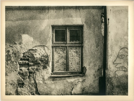 This is a lith print photograph depicting an old, weathered wall with a single window. The window has two upper panes and two lower ones, each covered with lace or floral-patterned curtains, partially obscuring the view inside. The wall itself is in a state of decay—plaster has crumbled away in several places, revealing the underlying brickwork. The surface is richly textured, with cracks, stains, and peeling layers contributing to a sense of age and abandonment.

To the right of the window, a simple line drawing of a face is visible, possibly graffiti. The expression appears stern or angry, adding a subtle narrative element to the scene.

The lith printing process is evident in the high-contrast tones, grainy texture, and warm, sepia-like coloration. Shadows are deep and rich, while highlights are soft and creamy, enhancing the tactile, almost painterly quality of the image. The border suggests it is a traditional darkroom print on old expired paper.