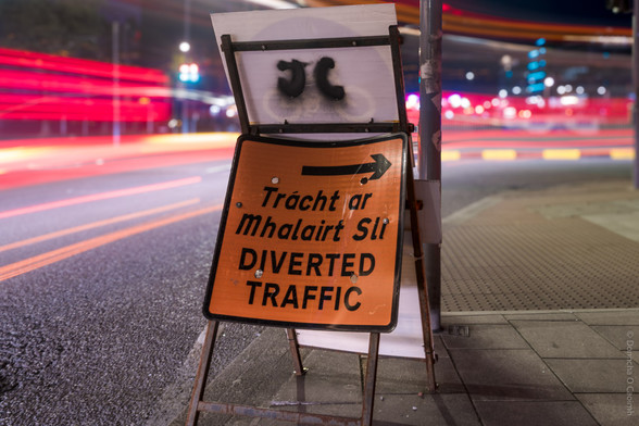 A nighttime street scene in Cork, Ireland, featuring a temporary orange road sign on a metal frame. The sign reads: "Trácht ar Mhalairt Slí DIVERTED TRAFFIC" with a black arrow pointing to the right. Behind the sign, blurred red and white light trails from passing vehicles are visible, and the street is illuminated by city lights.