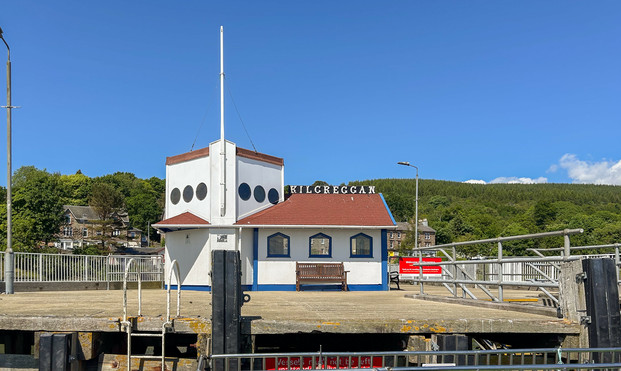 View of the Kilcreggan Calmac ferry terminal with a blue sky behind. The shot has been taken from the boat, so is looking up at the terminal building, with part of the wharf in the foreground. A number of saturated, contrasting colours are evident in the photo: greens of the trees in the background; the white terminal building with blue trims and a red slate roof; and the deep red of a Calmac sign.