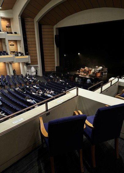 Photo of two chairs in a concert hall overlooking the auditorium. A complex setup of musical instruments is visible on stage, featuring a very large number of keyboards of various types, and including various pieces of digital audio equipment.