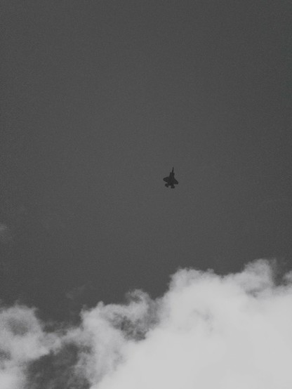 A black and white photo of a silhouette of a fighter jet flying high above white clouds against a muted gray sky.