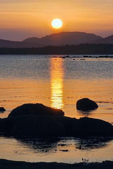 A vibrant sunset over the Atlantic Ocean on the west coast of Ireland, with the sun reflecting on the water and silhouetted rocks in the foreground, against a backdrop of distant hills.