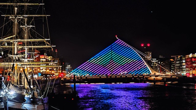 A nighttime view of the Samuel Beckett Bridge in Dublin, illuminated with vibrant blue and purple lights, next to a tall ship docked on the River Liffey. The city skyline with various buildings and their lights is visible in the background under a dark sky.