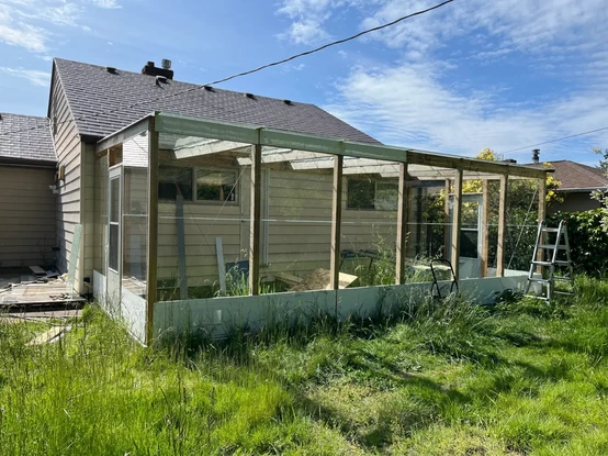 View of a wood and glass greenhouse attached to a home. The lawn is unkempt.
