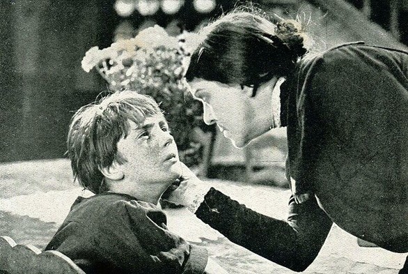 We can see a boy and a woman. The boy has short, straight, blondish hair and seems to be in distress. A woman is talking to him and touching his face with her right hand. She has long, tied, black, straight hair and is wearing a long-sleeved, black dress.