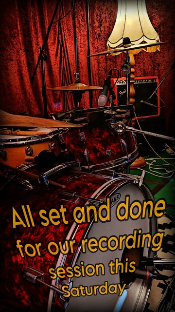 Zu sehen sind einige Mikrofone, ein rotes Drumset mit Bassdrum im Vordergrund, im Hintergrund sieht man eine alte Stehlampe und einen roten Fender-Amp. Im Hintergrund ein bordeauxfarbener Samtvorhang.