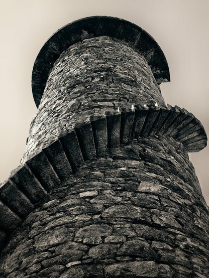 A low-angle, black and white shot of the Ballycorus Lead Mines chimney stack, showing its circular stone construction and the remnants of a winding stone staircase curving up its side. The tower tapers slightly towards the top, with a dark, circular cap.