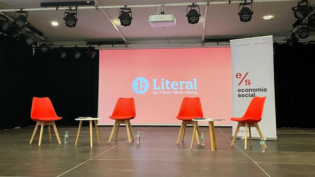 Un escenari amb dos grups de dos cadires vermelles i una tauleta baixa. Per darrera una projecció amb el logo de la Fira Literal, també en vermell.