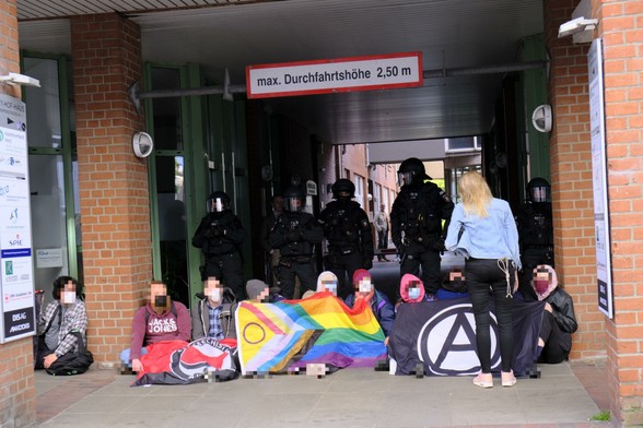 Sitzblockade am Zugang zur AfD Veranstaltung. Eine Person die zur Veranstaltung möchte steht vor der Sitzblockade. Im Hintergrund sind mehrere Polizist:innen zu sehen.