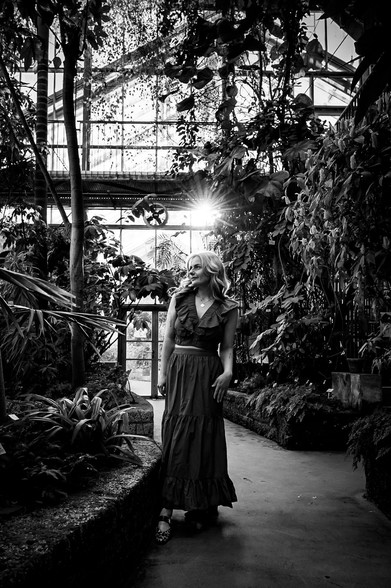 The black and white image captures a young woman, standing alone in a lush botanical garden. She as wearing a long dress and is posing, looking to the left. The sun behind her shines brightly through the glass panes of the structure. The foreground is filled with verdant plants, while in the background, the intricate architecture of the greenhouse is visible.