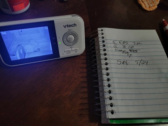 A baby monitor showing the back of a sleeping baby, next to a small notebook. Notes are:

f 6:05 3 oz
d 8:10
sleep 8:45 (struck out) 9p

New line
Sat 5/24