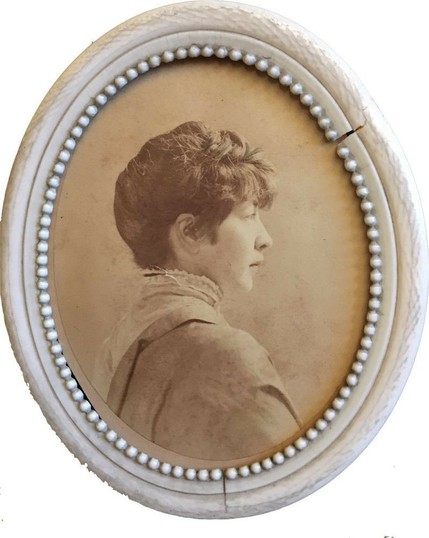 A sepia photograph of a young woman in profile, wearing late-nineteenth-century clothes. Her hair is worn up, above her ears, giving her a slightly boyish look. The photo is small, oval, and surrounded by a white frame; the frame is slightly cracked in two places.