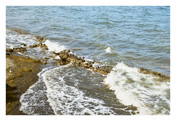 A slightly elevated, eye-level shot of a sea, meeting a rocky and somewhat muddy shore. The water appears blue-grey further out, transitioning to a murky brown near the shore where waves are breaking. - Google Gemini