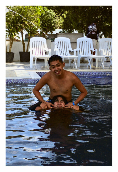 This full-shot photo captures a man and woman in a swimming pool on a bright day. The man, positioned behind the woman, has a wide smile and is shirtless. The woman is submerged up to her nose, with only her eyes and forehead visible. Her expression, with puffed cheeks and eyes looking up, suggests a playful interaction. Both appear to be having fun.

In the background, out of the water, several white plastic chairs are visible, along with some leafy green trees. A person wearing a black shirt and a dark cap is visible behind the chairs. The water in the pool is a dark blue, reflecting some of the surroundings. - Google Gemini