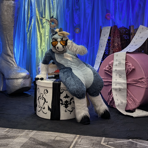 This is a photograph of a blue pegasus fursuiter posing in the Confuzzled photo room amongst props of oversized sewing kit and clothesmaking materials. He's got a bit of a lazy but fashionable slouch going on while wearing oversized orange aviator sunglasses, which he's tilting down so he can give the camera a nice judgemental stare.
