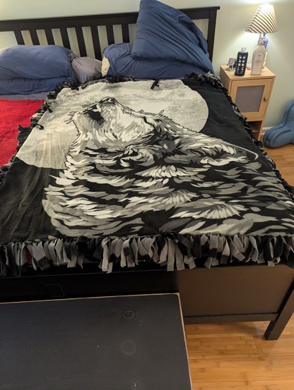 A fleece blanket with a tied fringe edge showing a very large and majestic wolf howling at a gigantic moon.