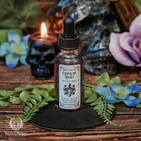 A clear glass vial sits on a black disc. The vial is filled with a clear-ish liquid and has an eyedropper in the screw-on cap. The label reads, "Bath & Body Oil / Cloak of Night / Dark Moon Magick / avocado oil, sunflower oil, jasmine essential oil, vetiver essential oil, fragrance oil, & mica. This is an exclusive item from Inked Goddess Creations.