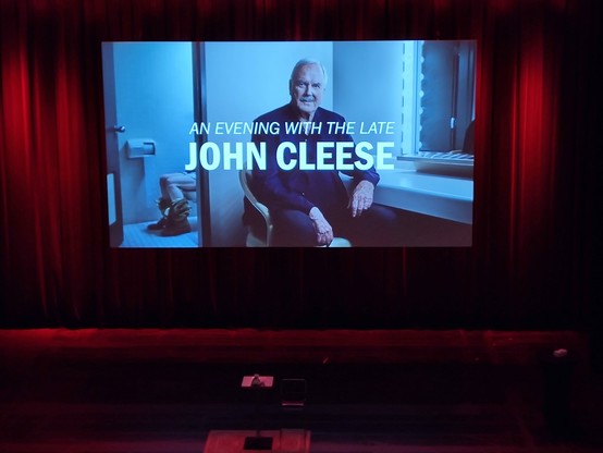 An empty stage with a still picture. Caption: an evening with the late John Cleese