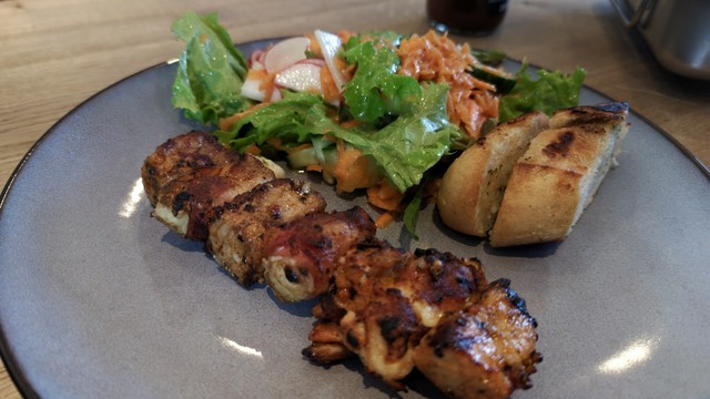 ein gegrillter "Gourmetspieß" mit etwas Kräuterbagutte und gemischtem Salat haben auf einem blaugrauen Teller mit dunklem Rand Platz genommen