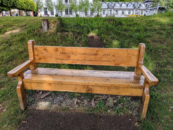 A wooden bench on grass with a white building in the distance. Carved into the seat back are the words "1944 ~ Brendan Clark ~ 2019". Underneath it says "What a time it was... Preserve your memories". The arm rests also have carvings. The left one (as we look at the bench) says "Old friends" and the right says "Bookends". All the words are from the song Bookends by Simon & Garfunkel. Presumably Brendan Clark, or whoever commissioned the bench, was a fan. Either way, it's a touching tribute to the deceased.