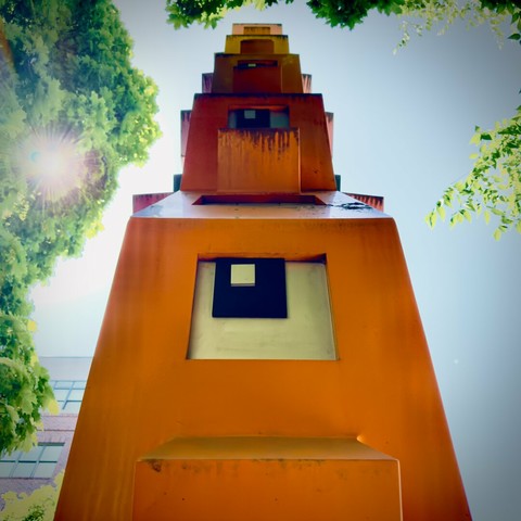 A large, orange, wooden totem pole stands tall under a city tree through which sunlight pierces on a clear day.
