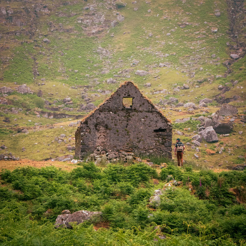 A man walks towards and abandoned stone structure in a green, rocky valley.