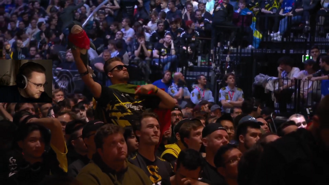 An enthusiastic esports fan wearing sunglasses and a team jersey holds up a blue IKEA Blåhaj shark plushie in a packed tournament arena, with crowds of spectators visible in the stadium seating behind him.