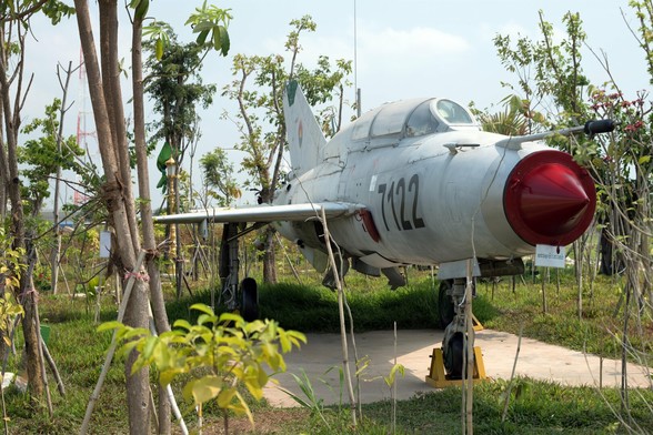 Photo shows a fighter jet placed on a concrete platform and surrounded by green trees and vegetation, slowly growing and envelop the weapons of war. It is a sunny day, and the sky is lightly clouded and light blue.