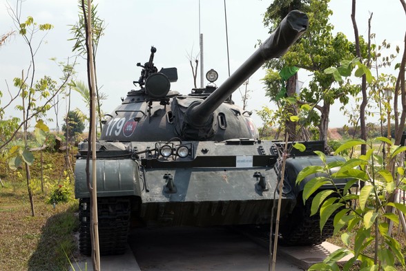 Photo shows an old main battle tank placed on a concrete platform and surrounded by green trees and vegetation, slowly growing and envelop the weapons of war. It is a sunny day, and the sky is lightly clouded and light blue.