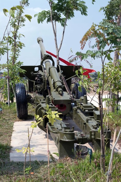 Photo shows a field canon placed on a concrete platform and surrounded by green trees and vegetation, slowly growing and envelop the weapons of war. It is a sunny day, and the sky is lightly clouded and light blue.