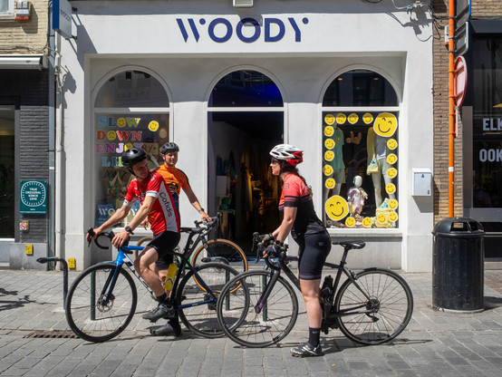 The image depicts three cyclists standing on a cobblestone street in front of a shop named "WOODY." The shop has a white facade with arched windows, and the name "WOODY" is prominently displayed above the entrance. The window displays colorful text that reads "SLOW DOWN ENJOY LIFE" and is adorned with yellow smiley face stickers.