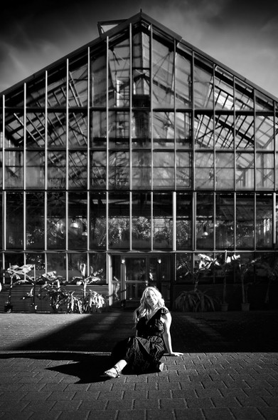 The image features a woman sitting in front of a large greenhouse. The greenhouse, constructed with glass and a metal frame, stands tall in the background. Plants are visible within the structure. The woman sits on a brick or stone-paved area in front of the building.