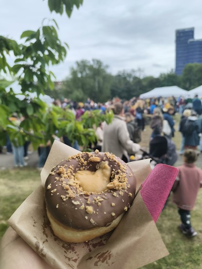 A hand holding a Snickers-flavoured donut, with a blurry outdoor scene and people in the background.
