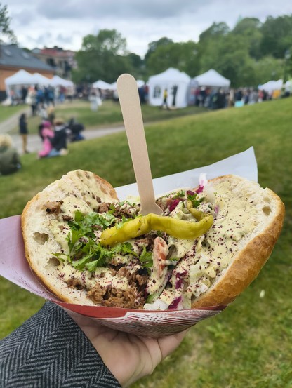 A person holding a large open-faced sandwich with vegan meat, vegetables, and sauce, with a blurry outdoor market in the background.