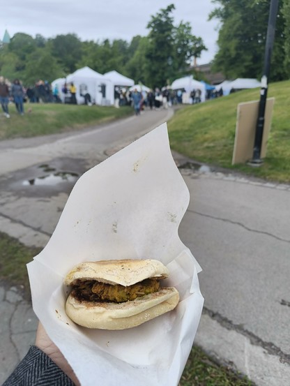A person holding an Indian vada pav   sandwich in a paper wrapper with a blurred outdoor market in the background.