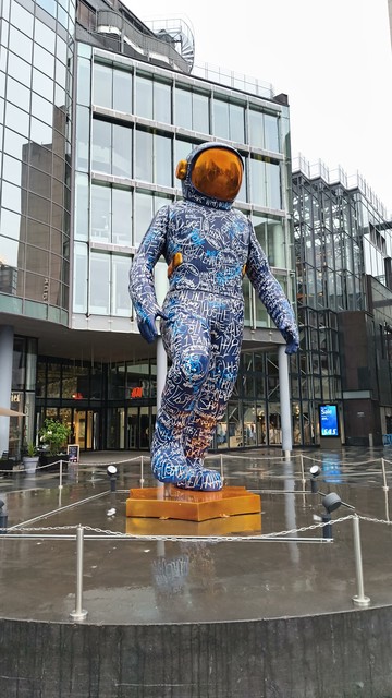 A large, colorful astronaut statue standing on a gold base in front of a modern glass building.