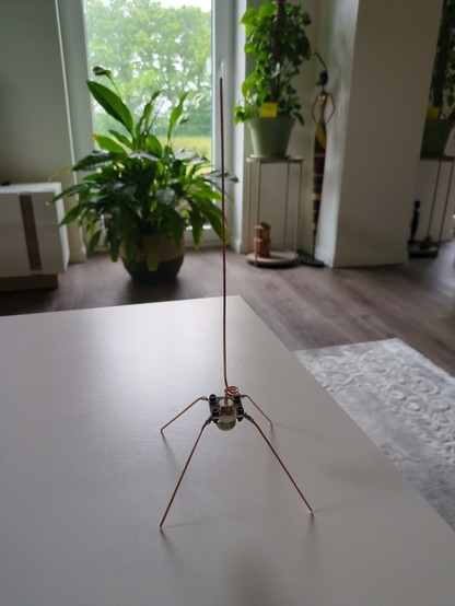 The 5/8 wave length ground plane antenna on top of a white table in the living room without any cables attached to it. 

It has 4 legs made of copper wire which are shaped like a pyramid. The legs are 82mm each. The top wire, the radiator, is pointing upwards and has a length of about 205mm. 

There are a window and some green plants in the background.